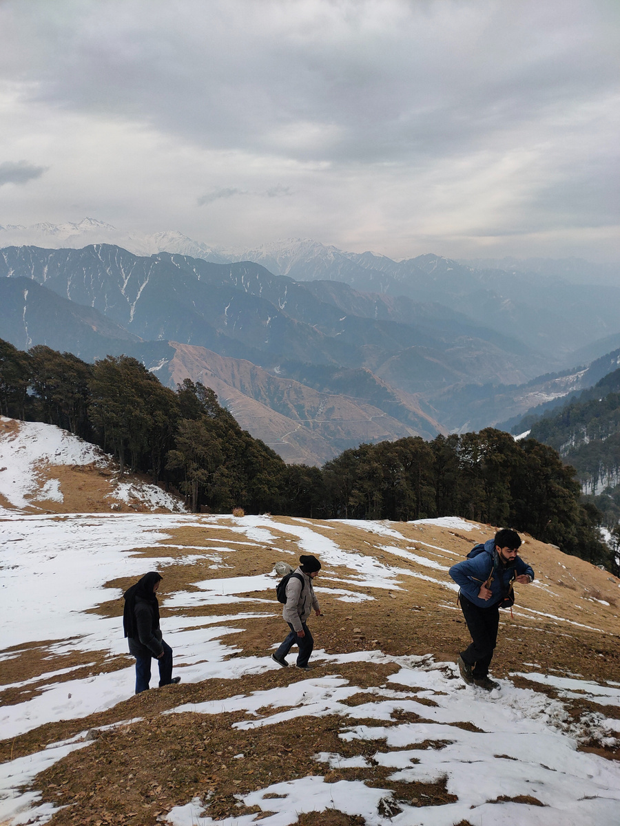 Hikers crossing a snowy ridge with the Dhauladhar range behind