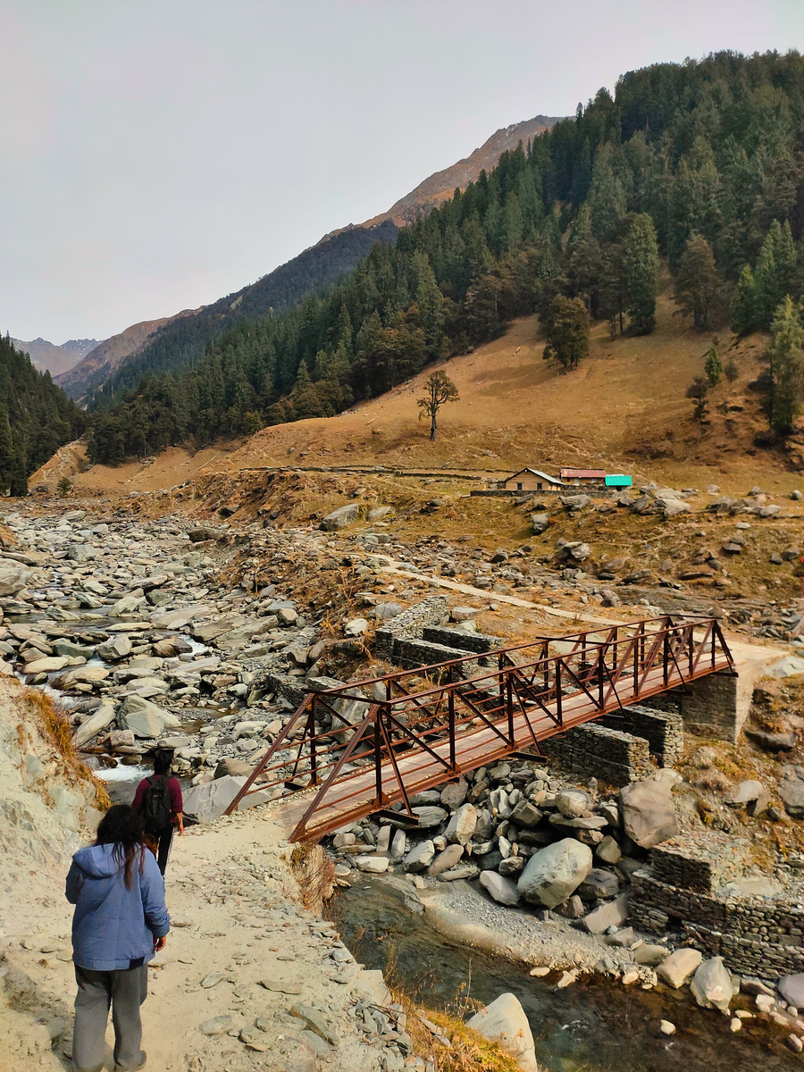 Hikers crossing a rugged mountain valley on a footbridge over a rocky river, Dhauladhar range behind